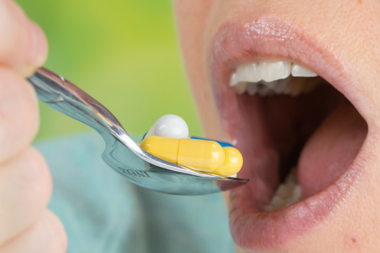 ihminen suu auki syömässä lusikalla keltaisia ja valkoisia pillereitä.